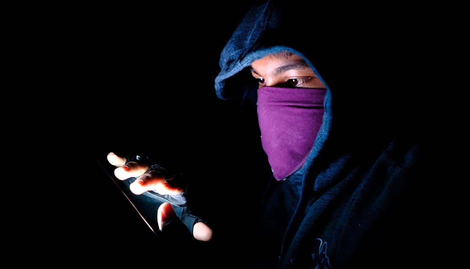 Image of hooded and masked man using a mobile phone signifying cyber espionage by hackers using fake apps with Android malware