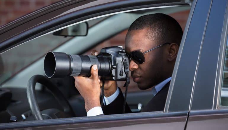 Image of man sitting in a car holding a camera with zoom lens signifying the use of online behavioral tracking for spying on users