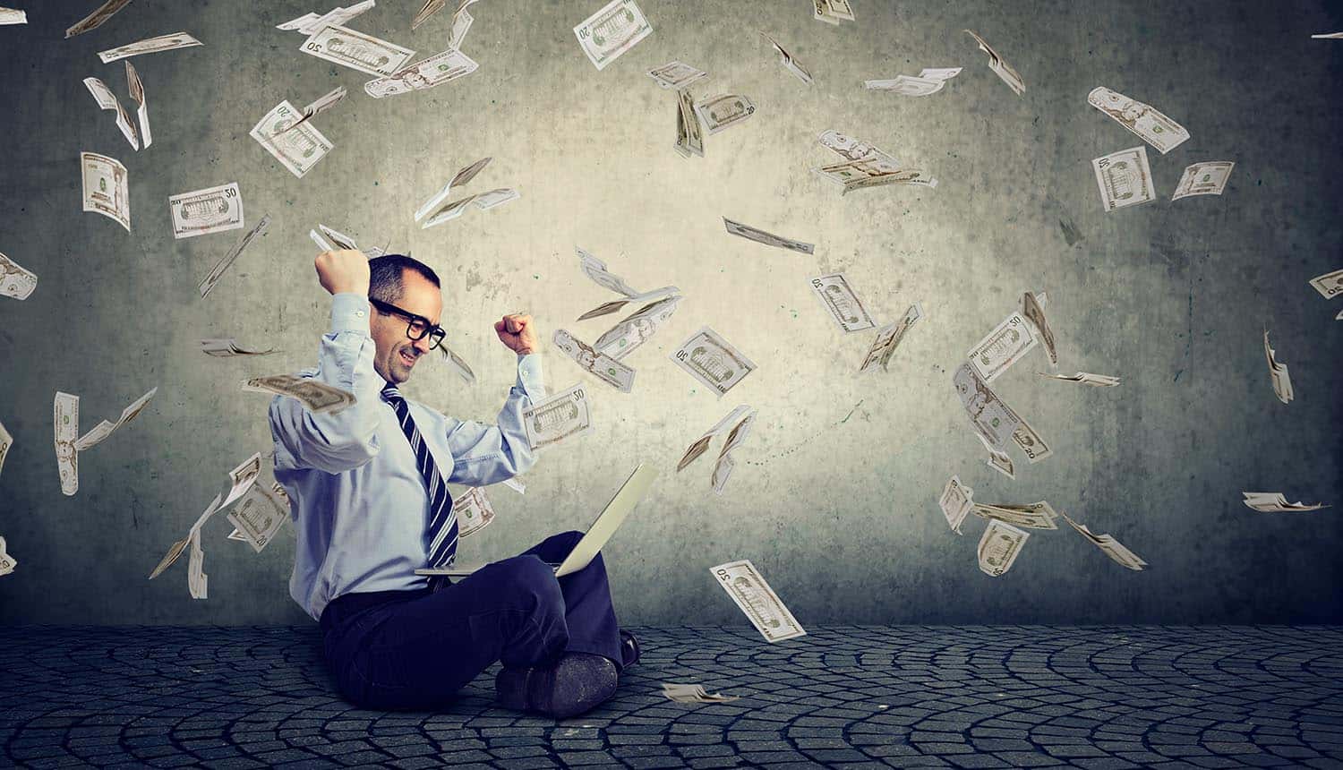 Image of man with arms raised sitting on floor with a laptop and cash flying around signifying the idea of how facebook will monetize your data