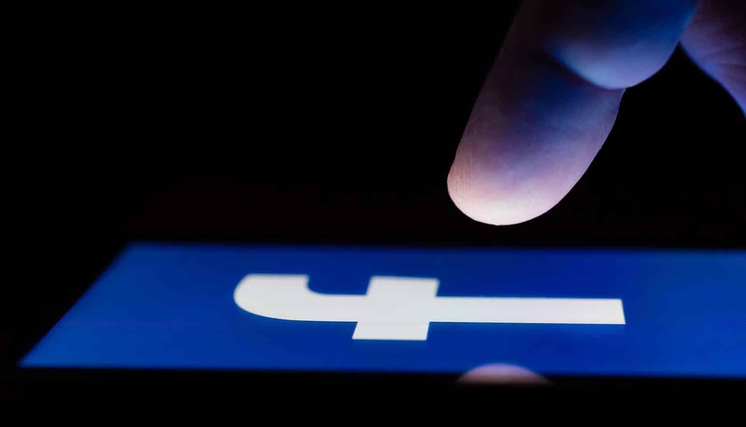 A smartphone lying on a table in the dark, displaying Facebook logo with finger above touch screen.