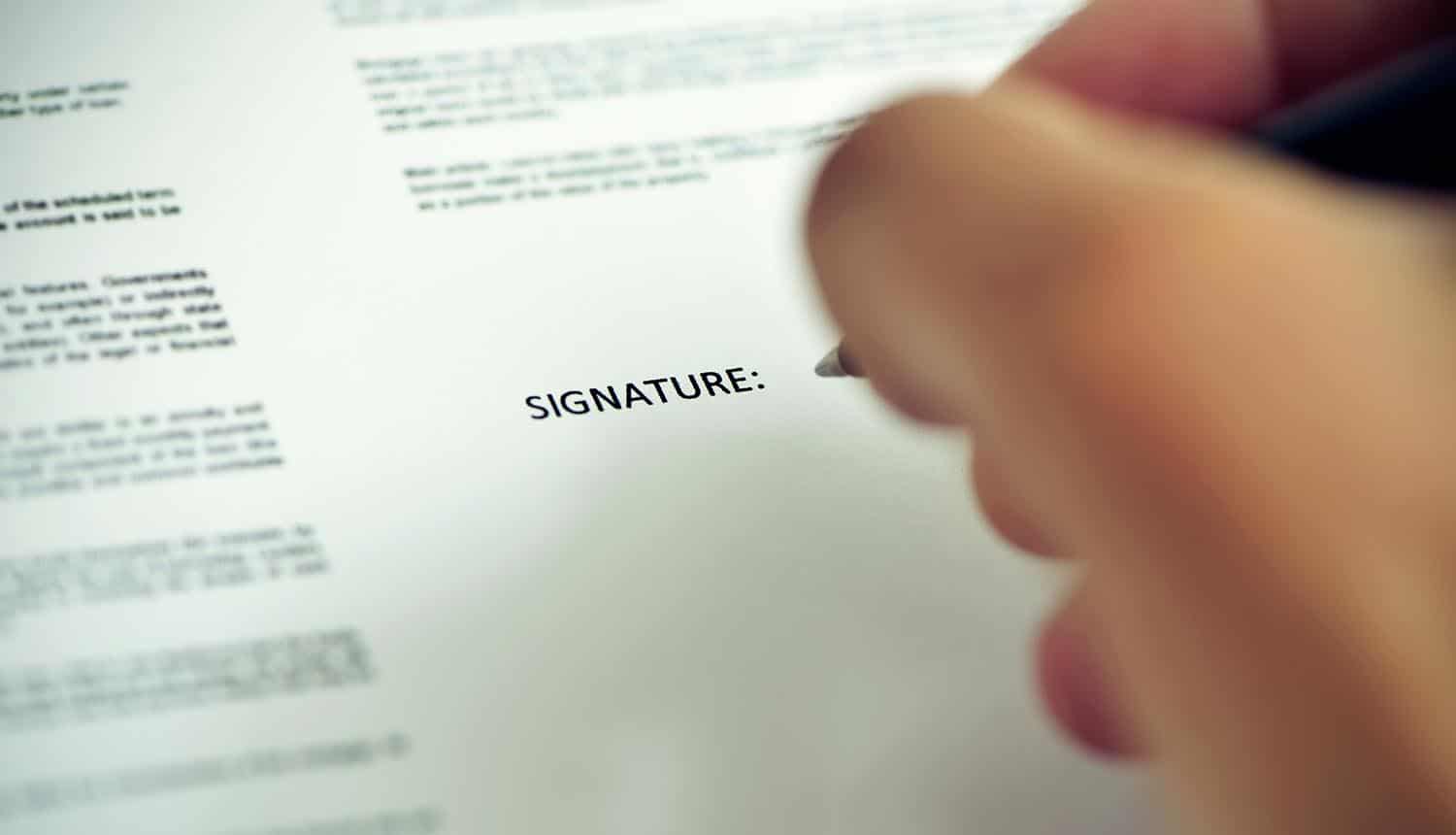 Closeup of a young man about to sign a document with a pen under the word signature showing marketing consent