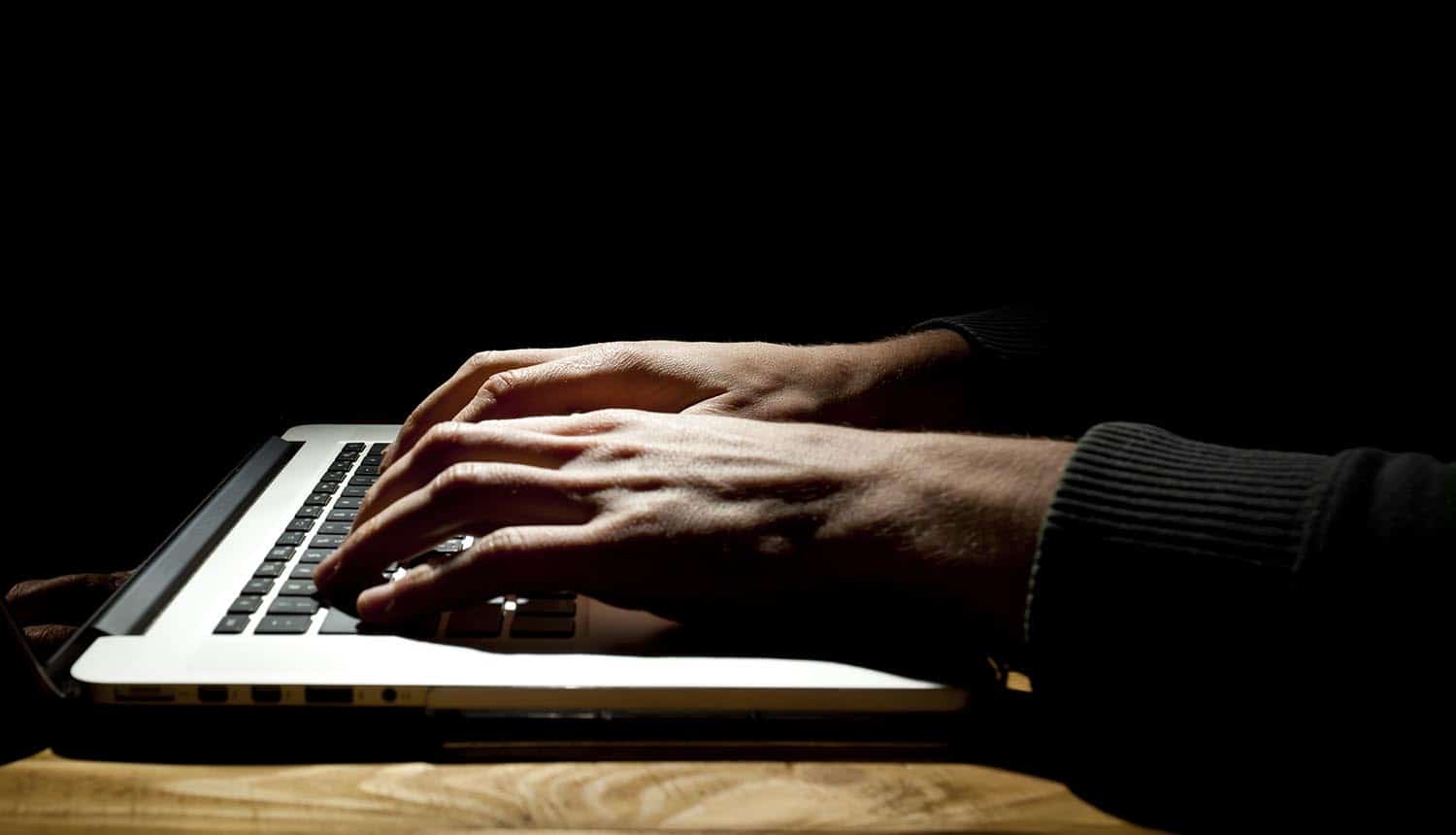 Man working on laptop in dark room showing increasing dark net hacking services targeted at enterprises