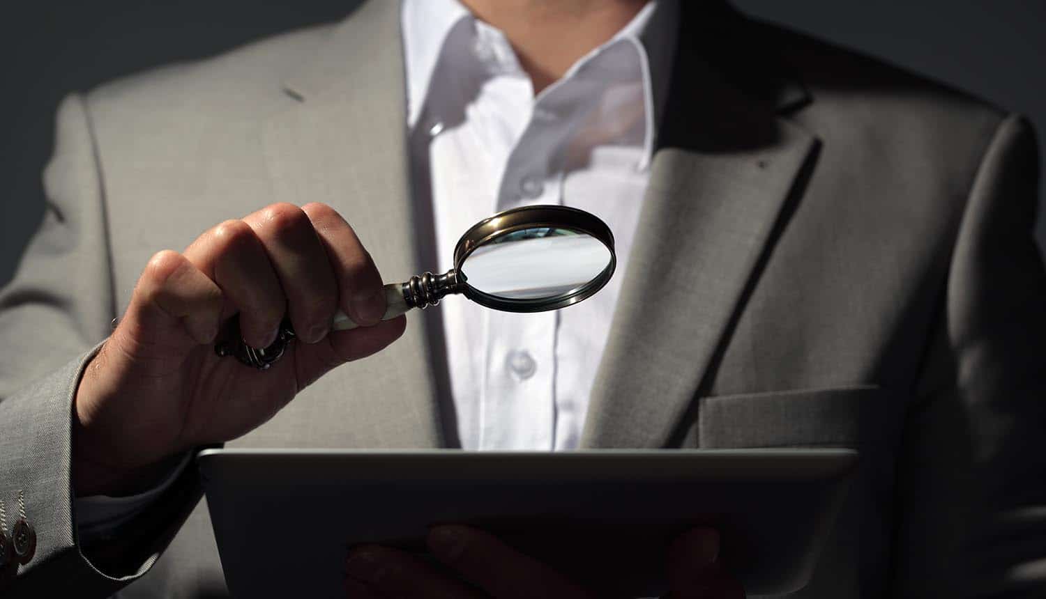 Businessman looking at tablet through magnifying glass showing the role of investor in boosting global cyber security as outlined in new WEF report