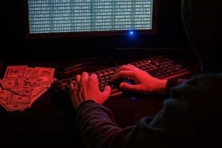 Man in hood working on computer next to pile of cash showing the financial impact of phishing scam